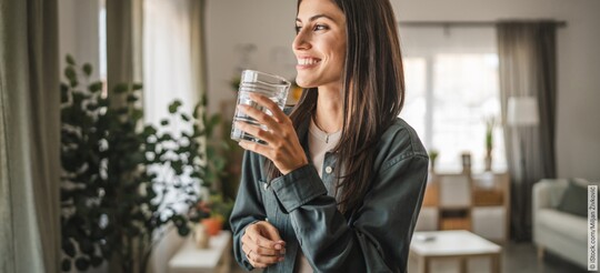 Eine lächelnde junge Frau steht in einem hellen Wohnzimmer und hält ein Glas Wasser in der Hand.