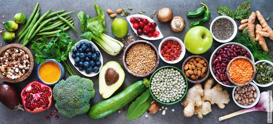 Eine farbenfrohe Auswahl an gesunden Lebensmitteln, darunter Brokkoli, Avocado, Beeren, Nüsse, Hülsenfrüchte und verschiedene Gewürze auf einem dunklen Untergrund.