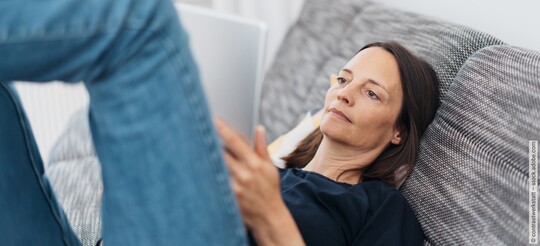 Frau informiert sich auf dem Sofa liegend mit ihrem Tablet.