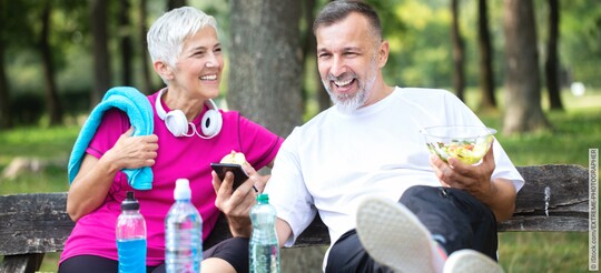 Zwei ältere Menschen sitzen gemeinsam nach dem Sport mit Essen auf einer Parkbank und blicken lächelnd auf ein Handy. 