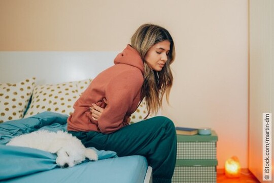 Junge Frau sitzt auf dem Bett und hat sichtlich Schmerzen wegen ihrer Blasenentzündung