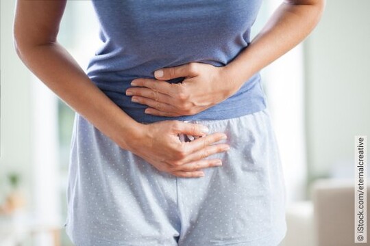 Frau im blauen Schlafanzugoutfit hält ihre Hände an den Bauch und ist leicht gekrümmt - sie hat sichtlich Schmerzen durch ihren Chronisch-wiederkehrenden Harnwegsinfekt