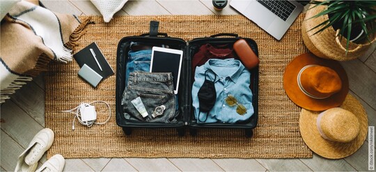 Offener schwarzer Rollkoffer auf dem Boden, gefüllt mit Kleidung, elektronischen Geräten und Reiseaccessoires, umgeben von einem Laptop, Schuhen und Hüten.