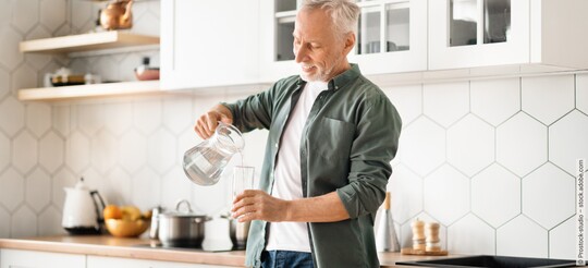 Ein älterer Mann steht in der Küche und gießt sich lächelnd ein Glas Wasser ein.