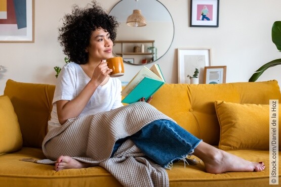 Eine junge Frau mit lockigem Haar sitzt auf einem leuchtend gelben Sofa, eingehüllt in eine gemütliche beige Decke, mit einem Buch in der einen und einer orangebraunen Tasse in der anderen Hand. Sie blickt nachdenklich in die Ferne, umgeben von einer stilvollen und pflanzenreichen Wohnumgebung. Diese ruhige Szene spiegelt entspannte Momente der Selbstfürsorge wider, die auch für das emotionale Wohlbefinden und die Blasengesundheit förderlich sind.
