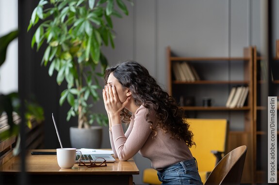 Eine junge Junge Frau sitzt am Schreibtisch vor ihrem Laptop und verbirgt das Gesicht in den Händen.
