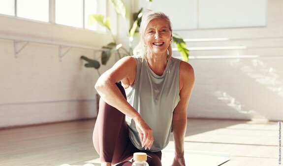 Eine Frau sitzt in sportlicher Kleidung auf einer Yogamatte und lacht in die Kamera.